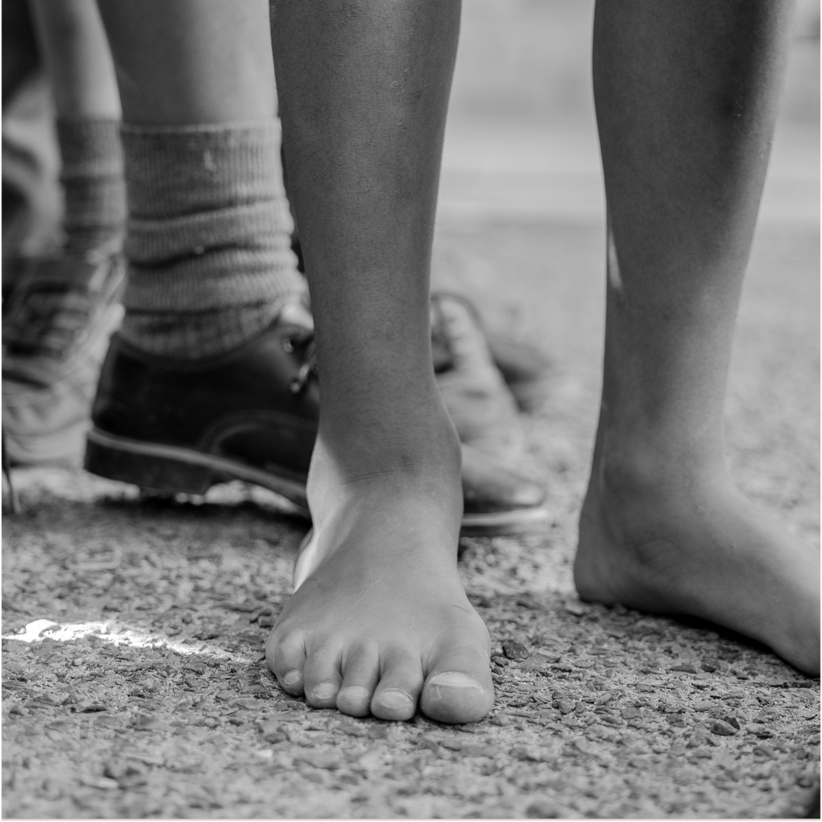 A learner standing barefoot on gravel beside classmates wearing school shoes and socks